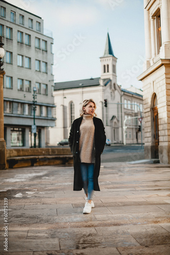 Stylish Woman in Black Coat and Blue Jeans Strolling Through Urban Landscape. Moody Cityscape with Fashionable Female Walking Along City Street. Urban Chic Concept with Woman Amidst City Buildings.