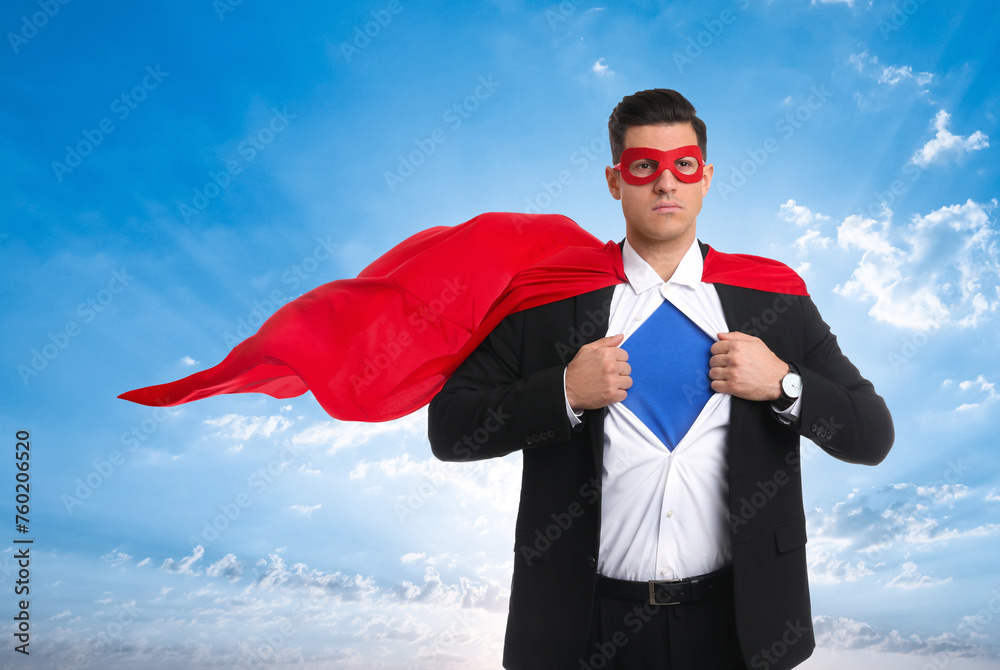 Confident man wearing superhero cape and mask taking suit off against blue sky with clouds Confident man wearing superhero cape and mask taking suit off against blue sky with clouds
