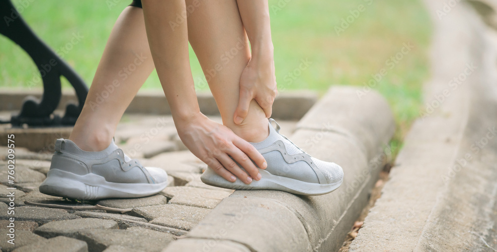 Sprained ankle problem. woman jogger. 30s asian female wearing sportswear holding her ankle with pain after running exercise in public park. - obrazy, fototapety, plakaty 