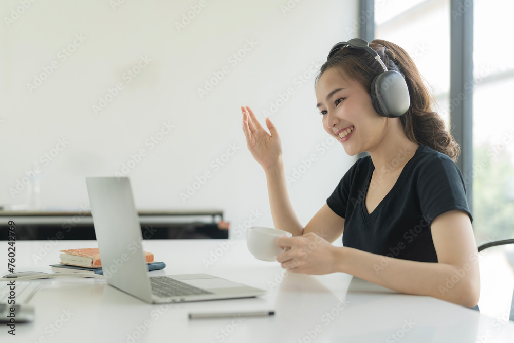 An Asian businesswoman wears headphones to meetings in video calls and drinking coffee at her home office.