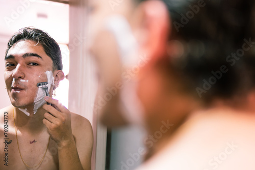Reflection of a joyful young Latino man engaging in his shaving routine in the bathroom mirror