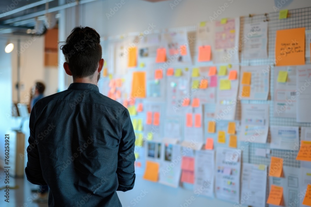 Man reviewing project plans on wall full of notes - A focused man viewed from behind analyzing a wall covered in project plans and colorful sticky notes Man reviewing project plans on wall full of notes - A focused man viewed from behind analyzing a wall covered in project plans and colorful sticky notes