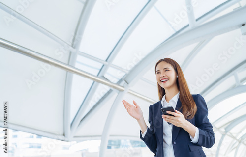 Smiling asian businesswoman holding smartphone while standing in front of modern business building, Concept of using the phone to communicate anywhere.