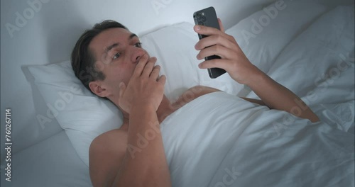 Man has been lying in bed since morning and, opening mobile phone, reads shocking news. Shocking headlines make us think about future. Shocking facts on screen make reconsider your plans for day.