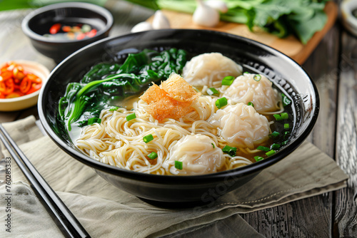 A steaming bowl of wonton noodle soup with fresh bok choy and a crispy garnish, on a wooden table.