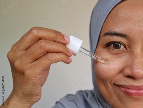 Half face of Happy muslim woman in hijab showing pipette with hydrating facial serum. Asian lady in headscarf applying serum on face with milia, dark sport, wrinkles and big pores.
