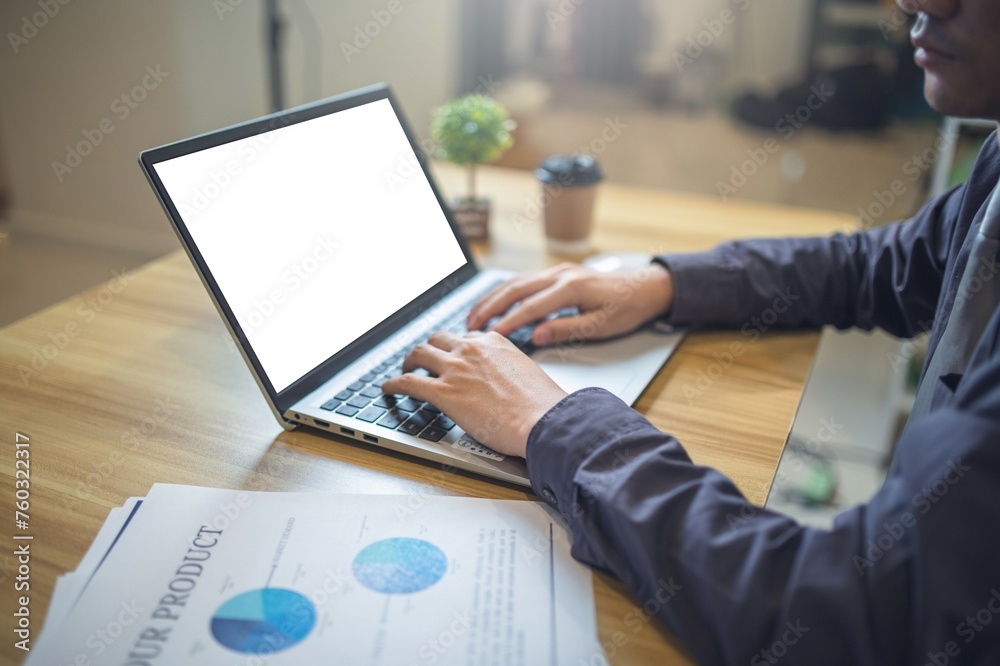 Business analyst reviewing pie charts on documents with a laptop with mockup screen on a wooden desk. Business analyst reviewing pie charts on documents with a laptop with mockup screen on a wooden desk.