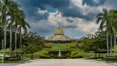 The beautiful presidential palace Istana Negara. A white building with golden domes and spires against a blue sky and clouds. In the foreground is a landscaped park with flower beds, a fountain.   photo