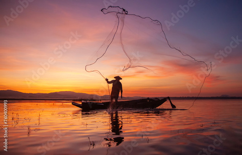 Fisherman casting his net on during sunset. Silhouette Asian fisherman on wooden boat casting a net for freshwater fish. Thailand culture.