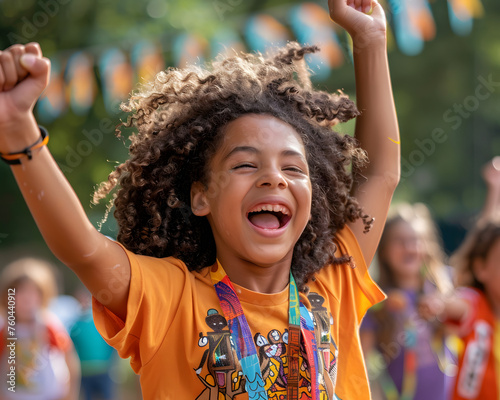 Exuberant young child with curly hair laughing and raising hands in victory at a fun outdoor summer camp celebration.