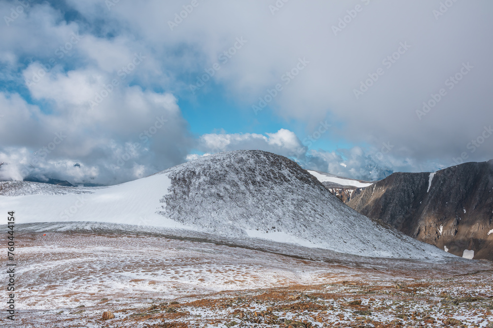 Awesome landscape with most beautiful rocky hill in freshly fallen snow among high mountains under cloudy sky. Colorful alpine view to snowy stony pass and wide snow-white glacier among low clouds.