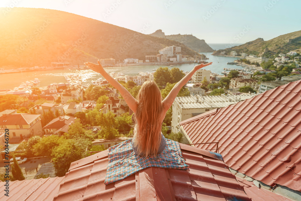 Woman sits on rooftop with outstretched arms, enjoys town view and sea mountains. Peaceful rooftop relaxation. Below her, there is a town with several boats visible in the water