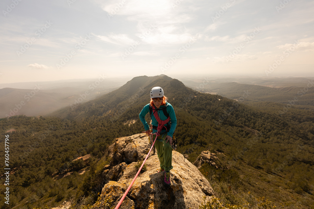 A woman is standing on a mountain top, wearing a helmet and harness. She is looking out over the landscape, taking in the view. Concept of adventure and excitement