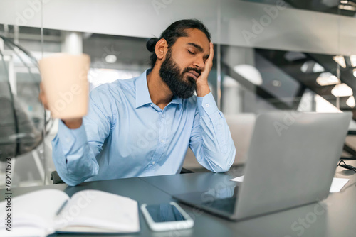 Sleepy indian guy office worker sitting at desk at office