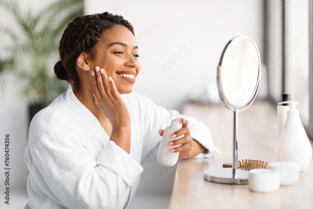 Smiling young black woman in white bathrobe applying skincare product Smiling young black woman in white bathrobe applying skincare product