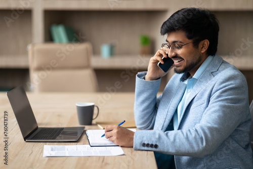 Middle Eastern businessman managing calls writing notes at office laptop