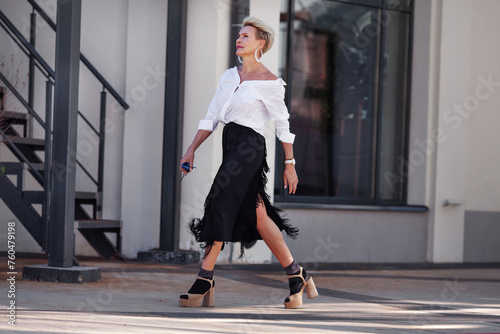 Confidence and sophistication mature woman walks city streets in fashionable ensemble chic white shirt and black fringed skirt with high heeled sandals and socks, fashion icon of style