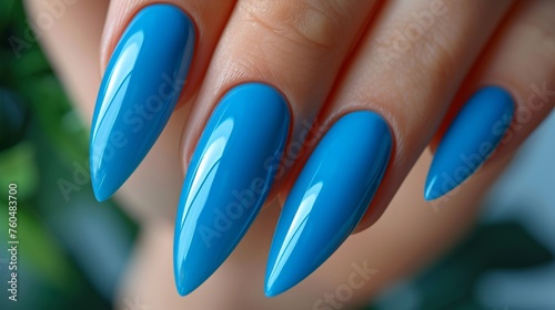 Elegant blue stiletto nails on a woman's hand with a green plant background, reflective of nature and style
