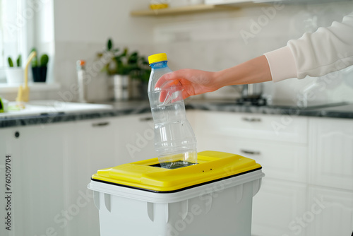 Woman hand leaving a plastic bottle in a plastic recycling bin with yellow lid in a white kitchen