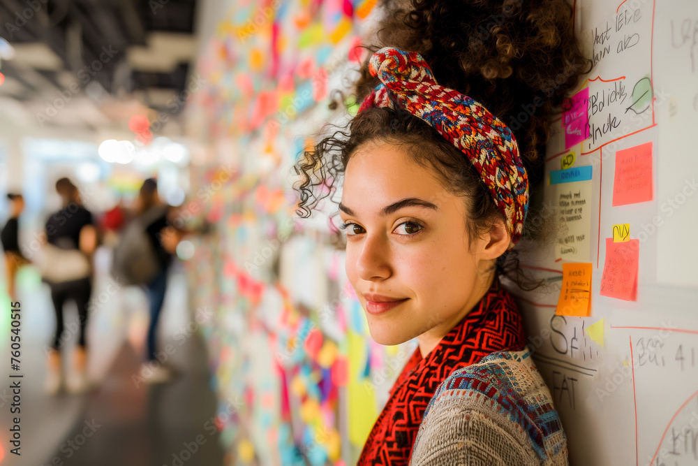 A young, curly-haired woman smiling in front of a colorful sticky note-filled board at a creative coworking space.
