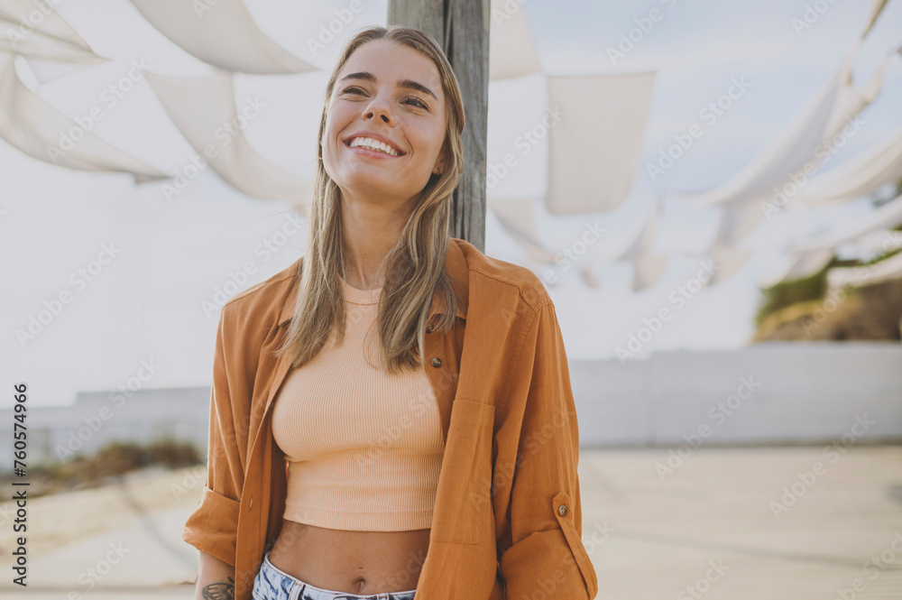 Young smiling cheerful fun cool minded calm woman she wearing orange shirt beige top casual clothes look aside in reverie mood rest relax walk outside in summer day. Urban lifestyle leisure concept. - obrazy, fototapety, plakaty 