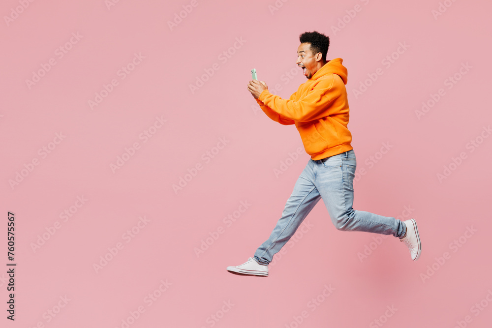 Full body side view young man of African American ethnicity wear yellow hoody casual clothes jump high hold in hand use mobile cell phone isolated on plain pastel pink background. Lifestyle concept