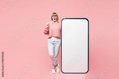 Full body elderly woman 50s years old wear sweater shirt casual clothes glasses big huge blank screen area mobile cell phone smartphone point index finger up isolated on plain pink background studio.
