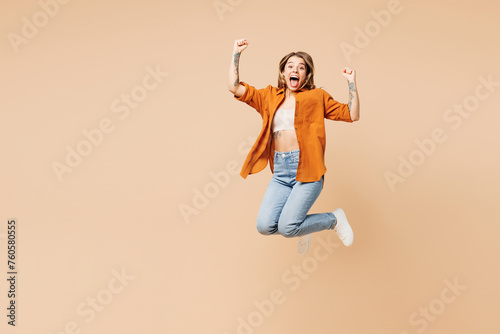 Full body cool young woman she wear orange shirt casual clothes jump high doing winner gesture look camera celebrate isolated on plain pastel light beige background studio portrait. Lifestyle concept.