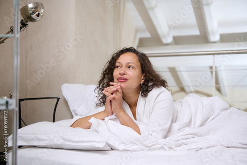 Happy positive woman smiling dreamily looking away, lying on the bed, enjoying her weekend morning while waking up