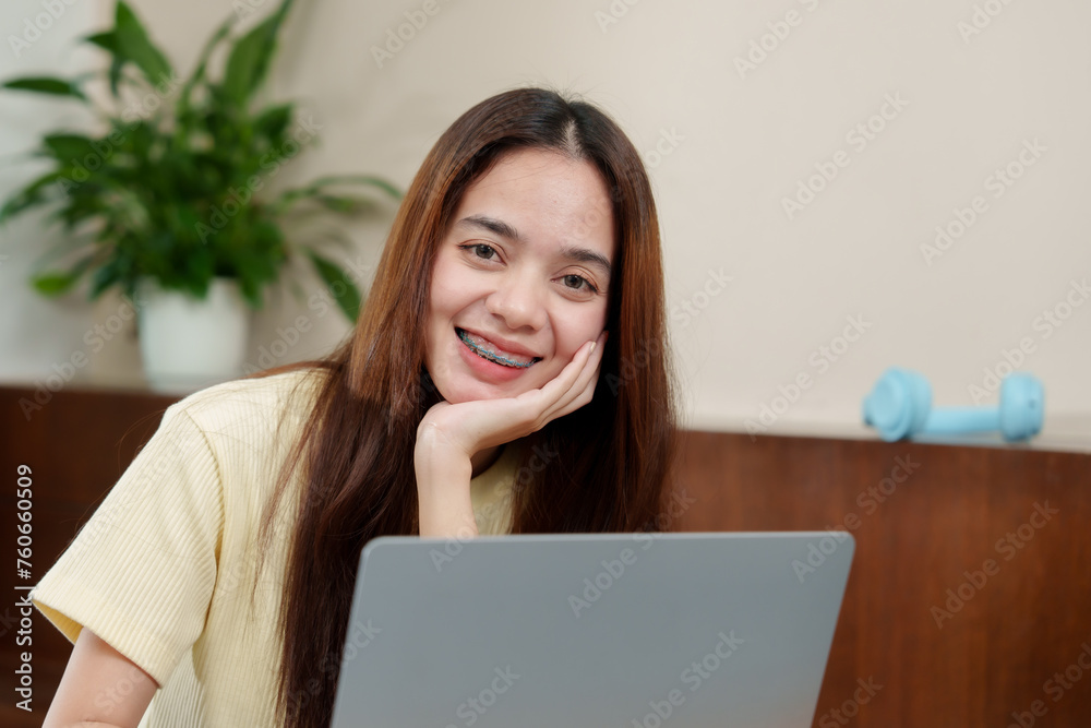 Joyful Asian woman with orthodontic braces looking at a laptop, hand on cheek, in a comfortable home setting. Cheerful person with a laptop beams while supporting face with hand, enjoying remote work Joyful Asian woman with orthodontic braces looking at a laptop, hand on cheek, in a comfortable home setting. Cheerful person with a laptop beams while supporting face with hand, enjoying remote work