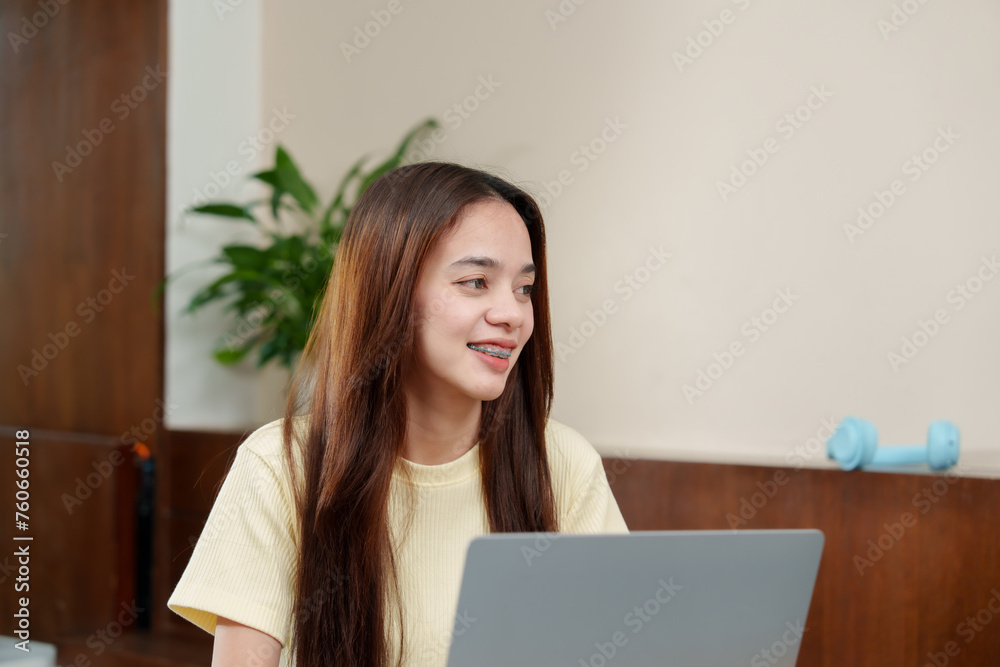 Young Asian woman with braces and long hair smiles gently while looking at a laptop screen in a well-lit room. Person with dental braces beams joyfully, eyes fixed on computer during casual e-meeting Young Asian woman with braces and long hair smiles gently while looking at a laptop screen in a well-lit room. Person with dental braces beams joyfully, eyes fixed on computer during casual e-meeting