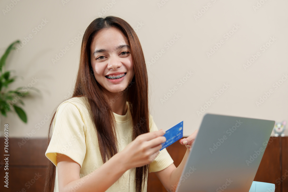 Jubilant Asian young woman with braces holds credit card, ready for online shopping spree, with laptop in front. Cheerful individual displays a bank card, preparing for e-commerce transaction, Jubilant Asian young woman with braces holds credit card, ready for online shopping spree, with laptop in front. Cheerful individual displays a bank card, preparing for e-commerce transaction,