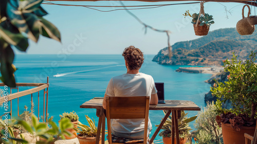 Rear view of a male freelancer working on his laptop, sitting at a table on a terrace overlooking the sea. photo