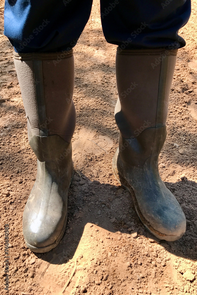 Close-up of dark green rubber boots of a farmer standing on clay soil on the road