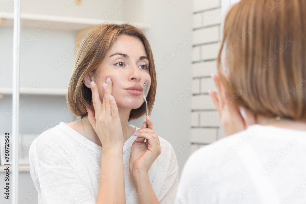 Attractive girl doing facial workout with spoon exercises in front of the mirror. Young woman holding spoon with mouth Attractive girl doing facial workout with spoon exercises in front of the mirror. Young woman holding spoon with mouth