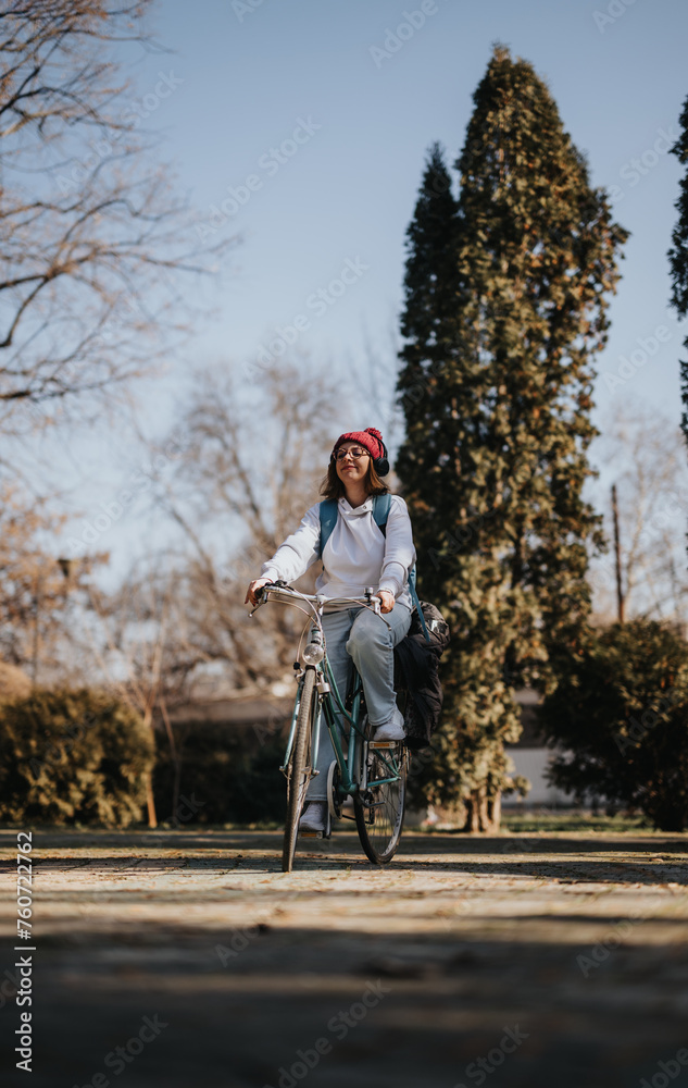 A woman with a red beanie and headphones smiles while cycling on a peaceful path amid tall trees, experiencing the beauty of winter.
