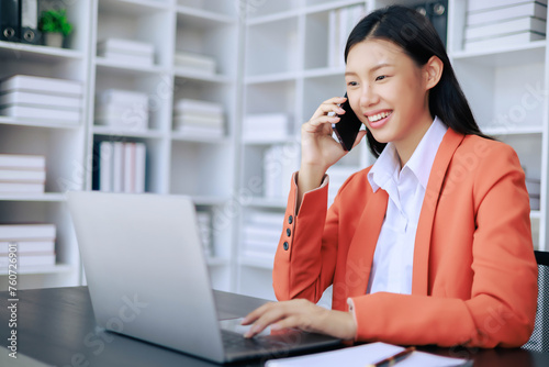 A woman is talking on her cell phone while sitting at a desk with a laptop