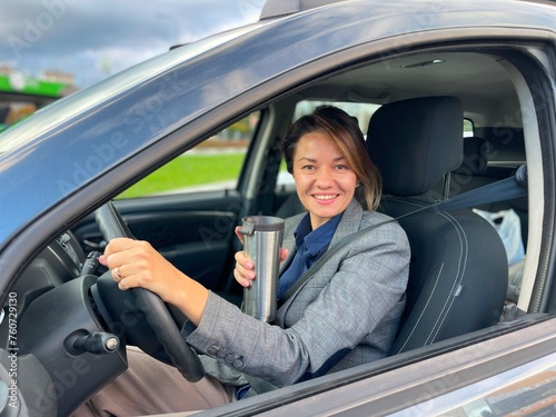 Cheerful Caucasian business lady sitting inside the car holding thermos with coffee looking at camera with smiled face. Businesswoman driving her vehicle and smiling. Close up © Nataliya