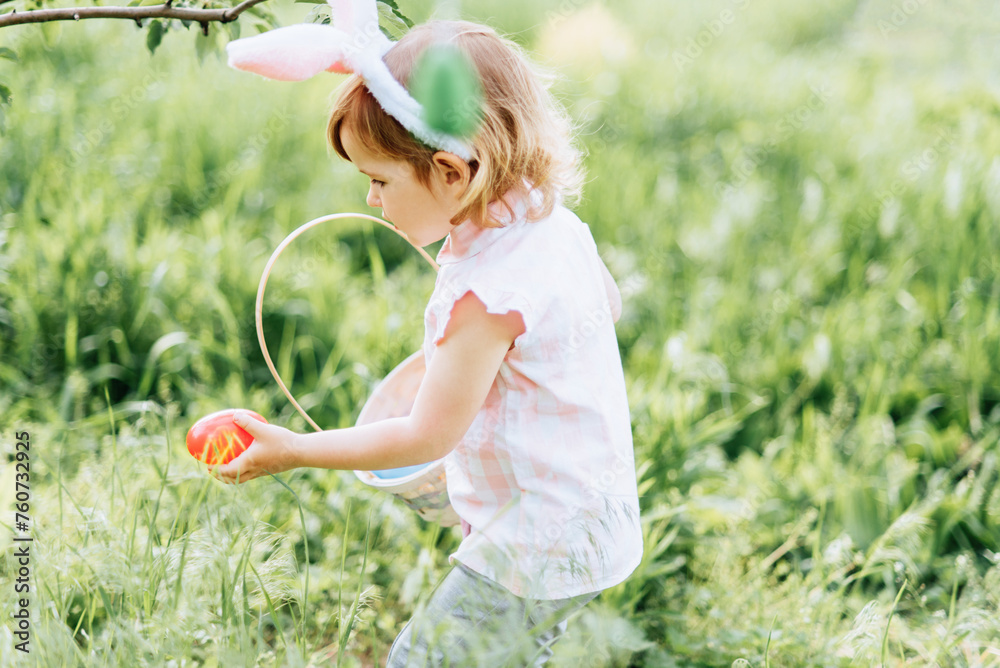 Easter egg hunt. Girl child Wearing Bunny Ears Running To Pick Up Egg In Garden. Easter tradition. Baby with basket full of colorful eggs. wide angle view