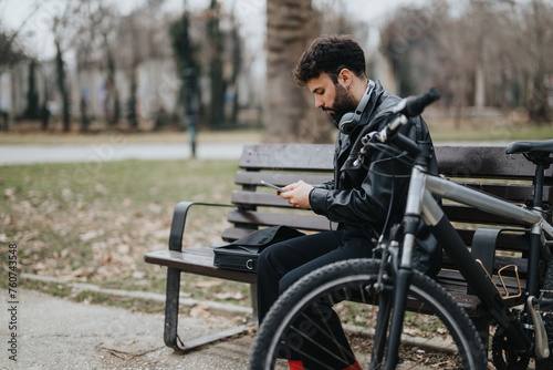 Handsome, stylish businessman enjoys the tranquility of nature while working outdoors with his smart phone, sitting on a bench beside his bicycle.