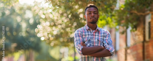 Serious young man in plaid shirt standing with arms crossed in a campus setting.