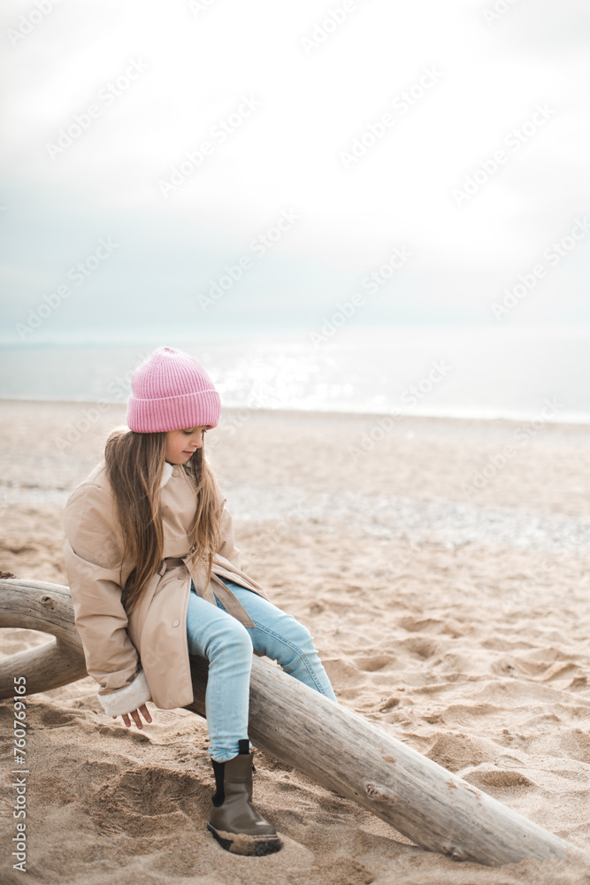 Stylish kid girl 5-6 year old wearing beige jacket sitting on dry tree over sea background outdoor. Spring season. Childhood.