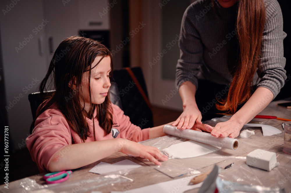 Intently focused, a mother assists her daughter with a clay craft project at home using rolling pin, showcasing a moment of shared concentration and educational interaction Intently focused, a mother assists her daughter with a clay craft project at home using rolling pin, showcasing a moment of shared concentration and educational interaction