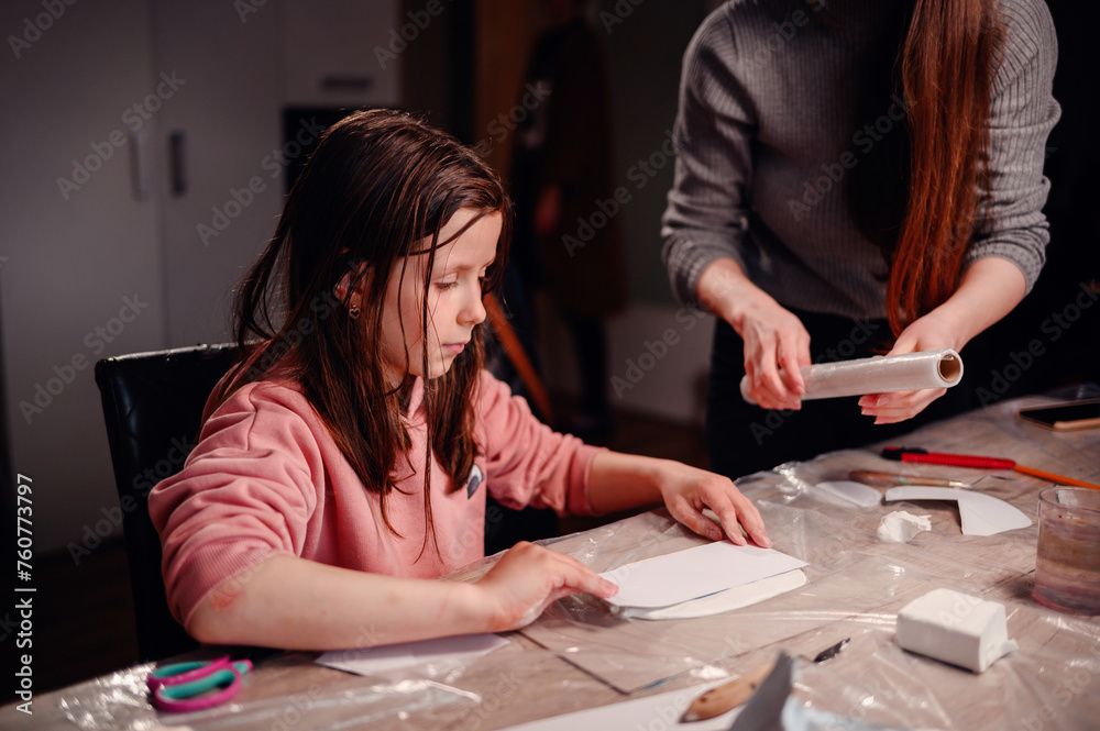 Fototapeta premium Intently focused, a mother assists her daughter with a clay craft project at home using rolling pin, showcasing a moment of shared concentration and educational interaction