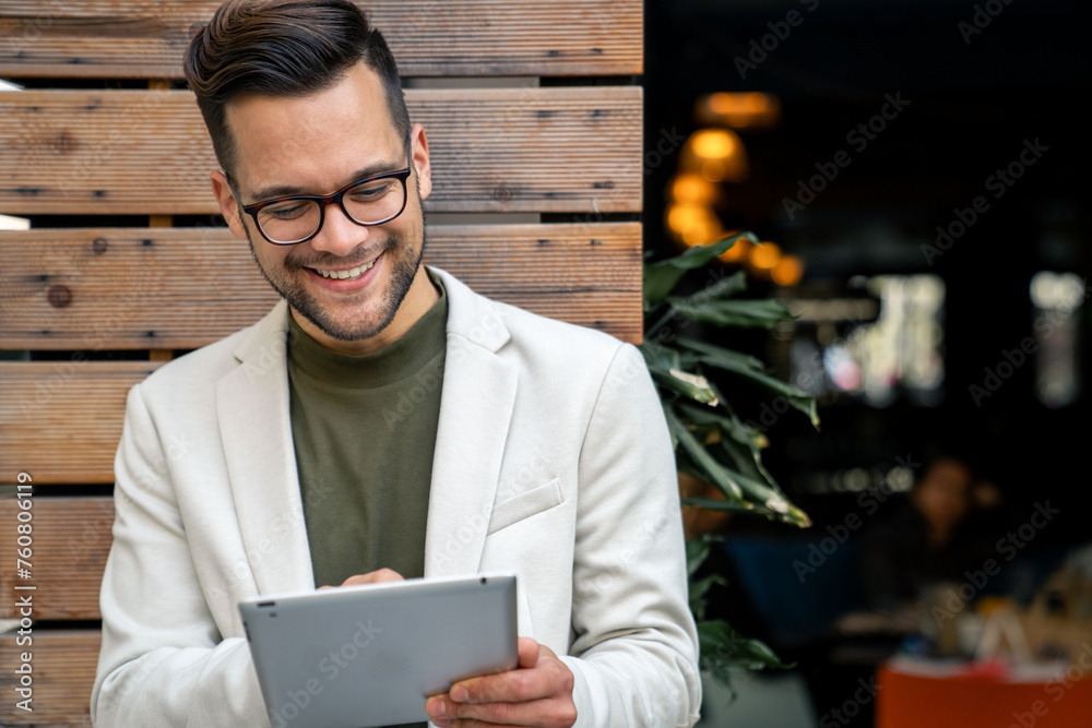 Handsome male business person standing outdoors in front of a local coffee shop and checking financial reports on his digital tablet, widely smiling.