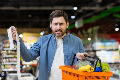 Portrait of unsatisfied shopper, expensive goods in supermarket, mature man looking upset at camera, with basket of groceries and huge check in hands. photo