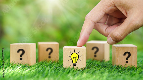 Hands point on yellow light bulb symbol on wooden cube with blurred question mark blocks in natural background for new idea concept, innovation power, creativity, Solution problem solving.