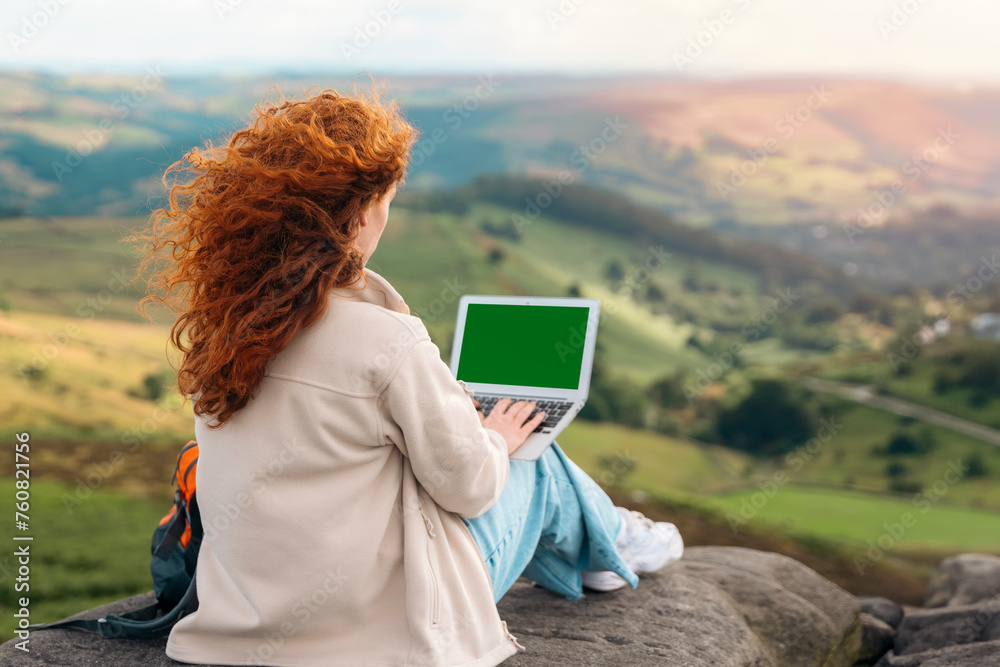 enjoying redhead woman reaching the destination, using laptop, and enjoying mountains landscape on the top of mountain at sunset. Enjoy hiking and exploring new places concept
