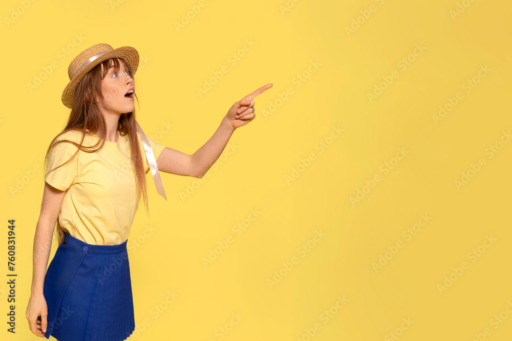 Surprised young redhead woman girl in a yellow t-shirt, blue skirt, and straw hat pointing her index fingers aside up on a yellow background with copy space. Emotions lifestyle concept Surprised young redhead woman girl in a yellow t-shirt, blue skirt, and straw hat pointing her index fingers aside up on a yellow background with copy space. Emotions lifestyle concept
