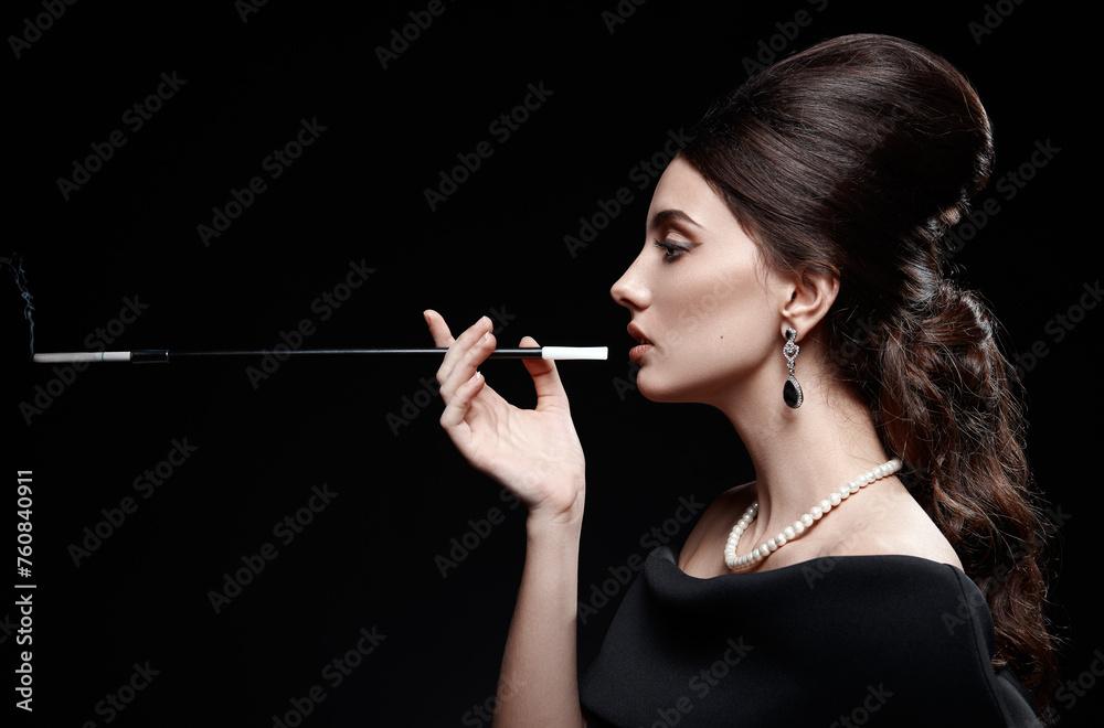 Obraz premium Retro shot: beautiful young woman smoking cigarette. Vintage portrait of pretty girl in 60s style. Gorgeous lady in black dress and pearl necklace against black background. Profile view